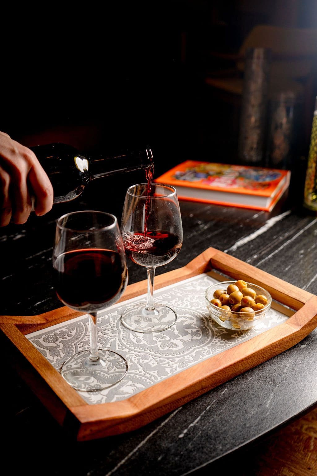 Red wine poured into glasses beside a bowl of nuts on a patterned serving tray
