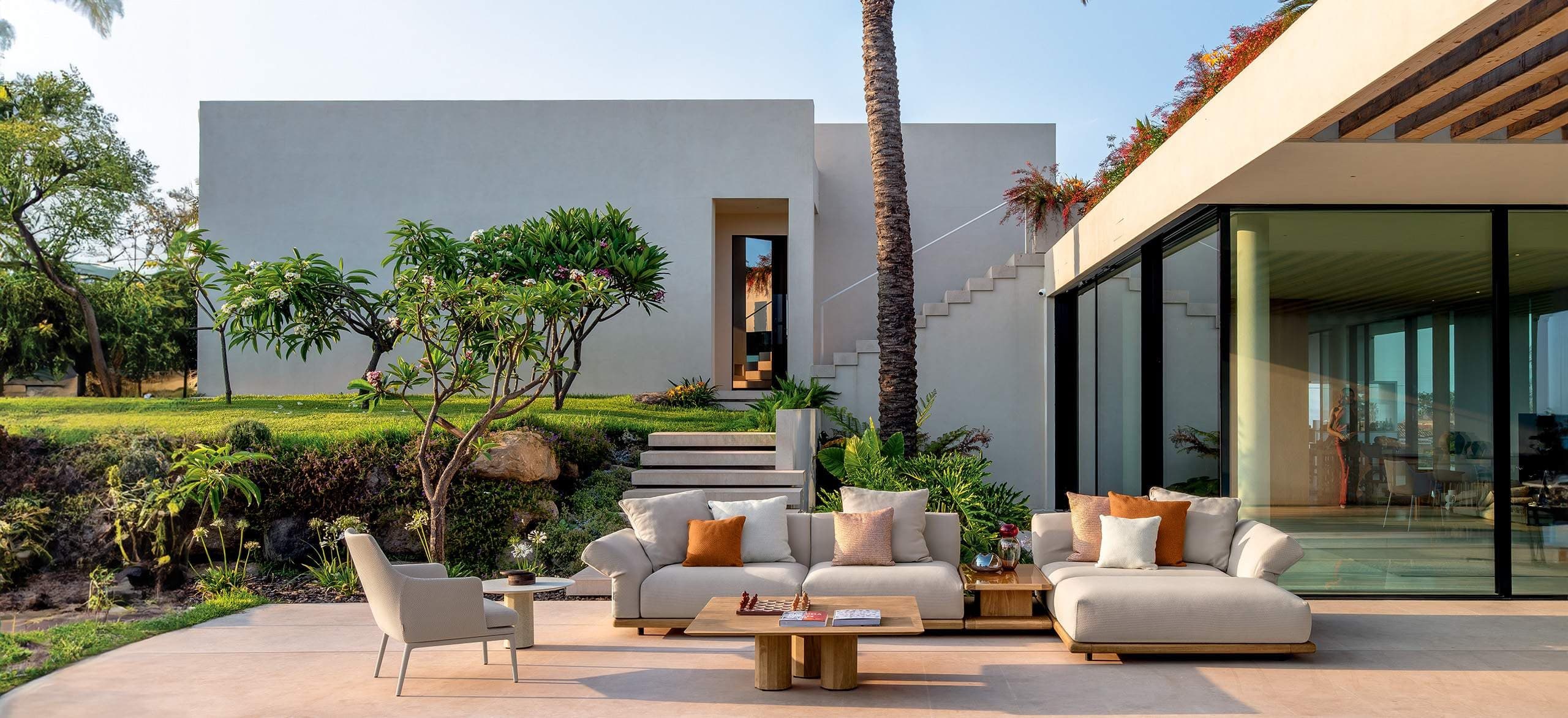 Luxury outdoor patio with cream sectional sofa, terracotta cushions, and wooden coffee table set against a modern white villa with lush tropical garden