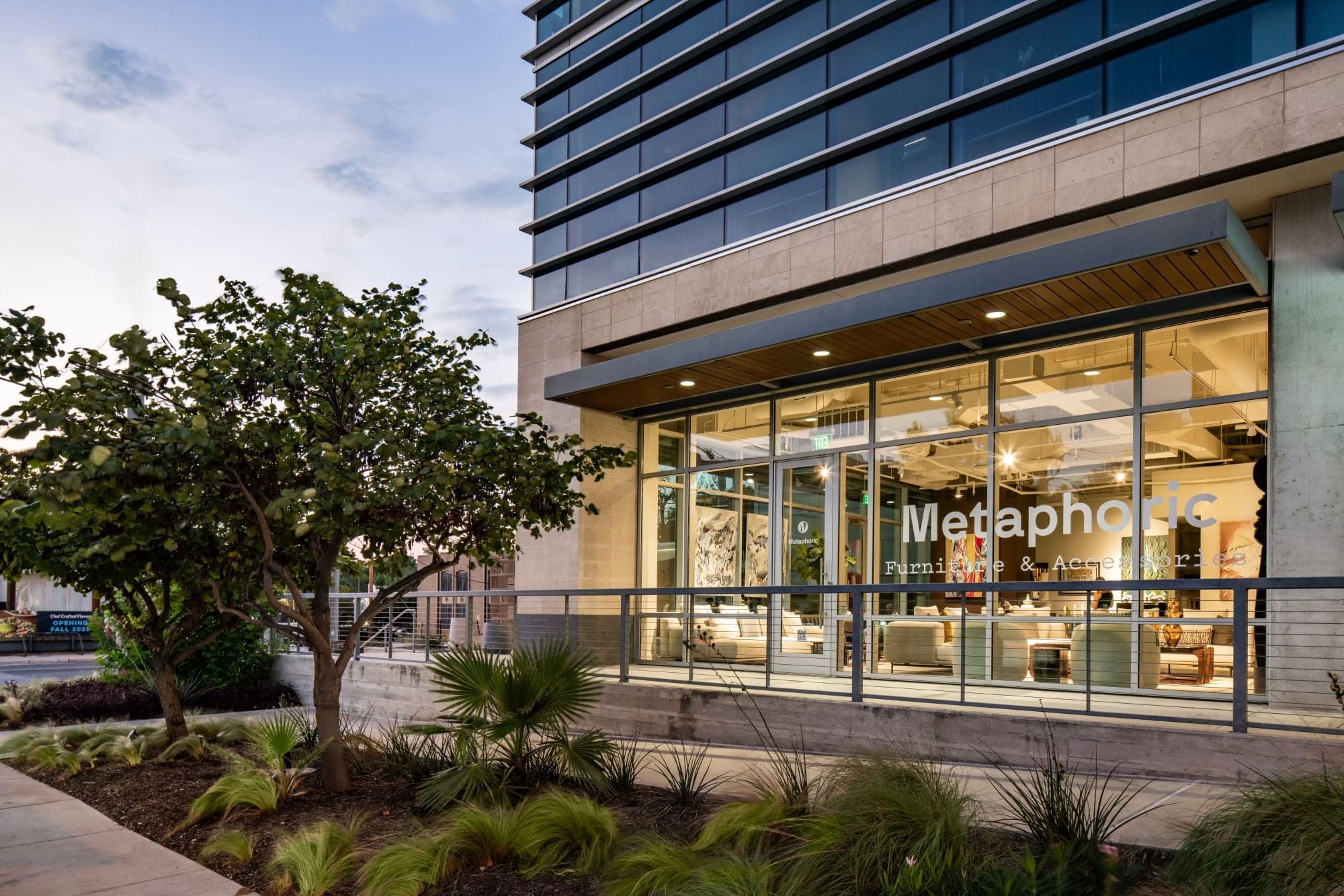 Metaphoric Furniture and Accessories storefront with floor-to-ceiling windows at ground level of a modern glass high-rise building at dusk