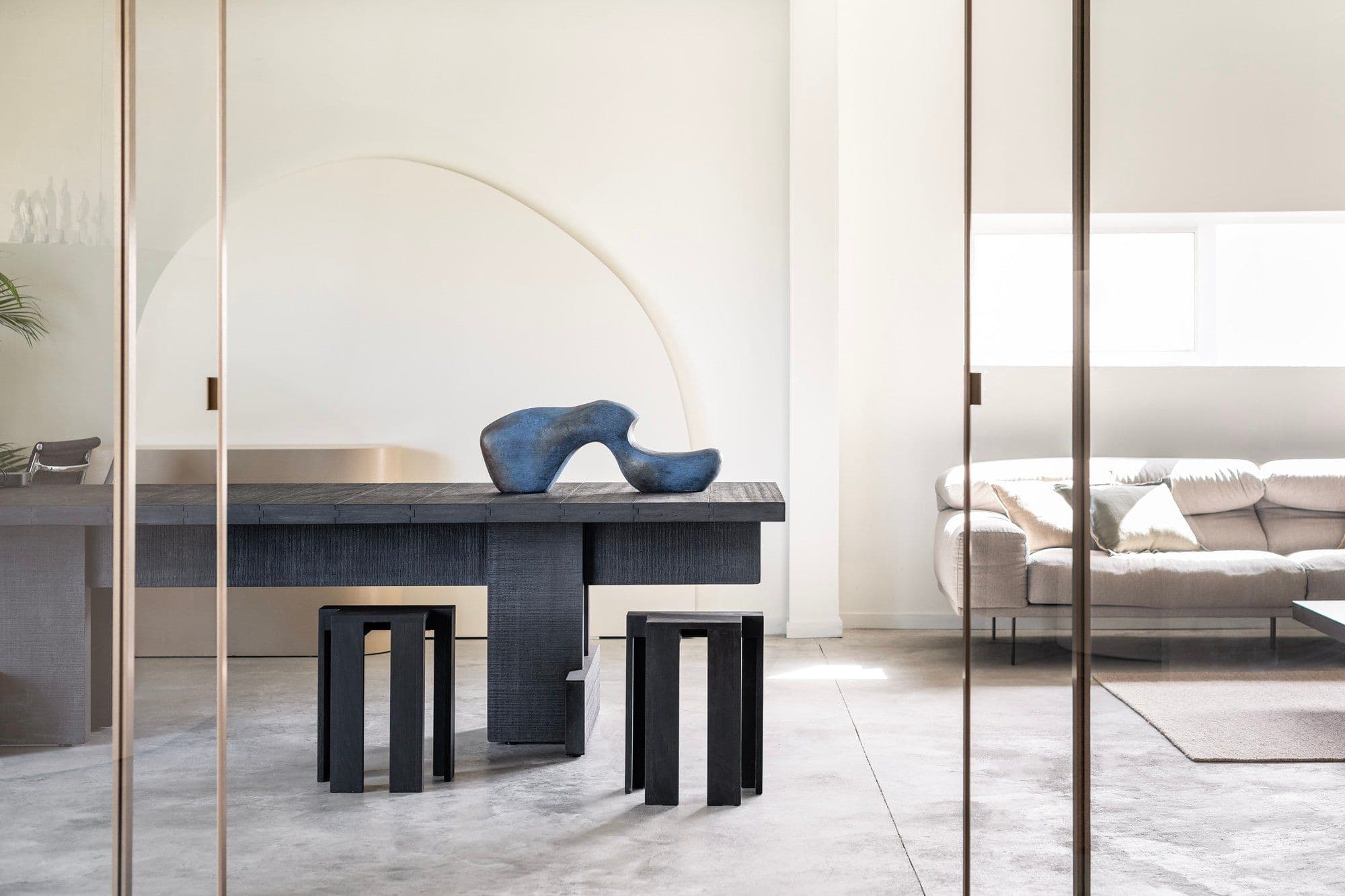 Dining room with a dark table and stools, a blue sculpture on the table and a pale arched wall behind glass doors