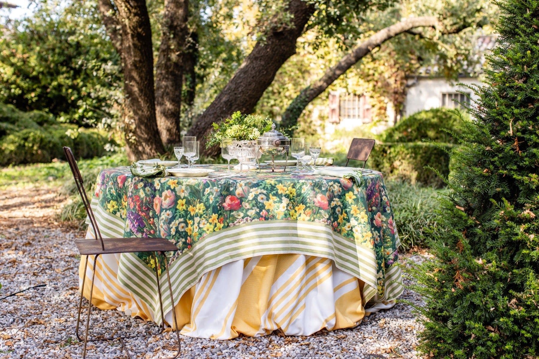 Elegant outdoor garden table set with floral and striped tablecloths, crystal glassware, and a silver centerpiece surrounded by lush trees
