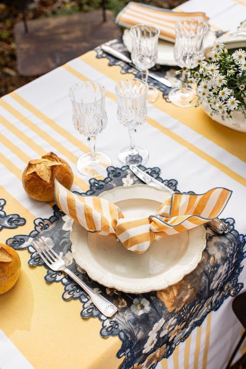 Striped yellow and white table runner with blue floral placemats, white shell-shaped plate, orange striped napkin, and crystal glasses in outdoor dining setting