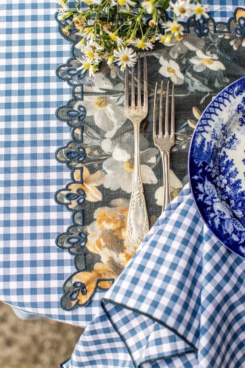 Blue and white gingham tablecloth with ornate floral placemat, silver forks, daisy flowers, and blue patterned plate styled flat lay