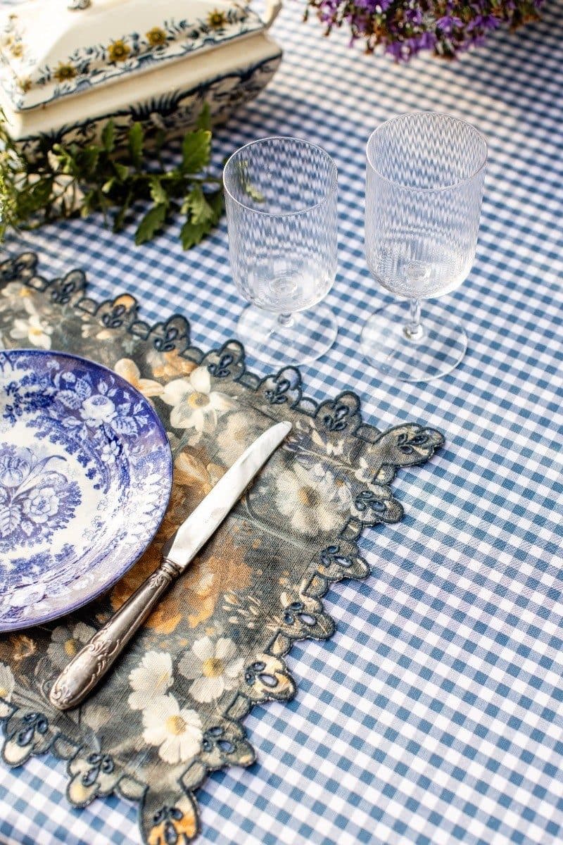 Blue and white gingham tablecloth with blue floral placemat, patterned napkin, decorative plate, knife, and two textured water glasses in outdoor dining setting